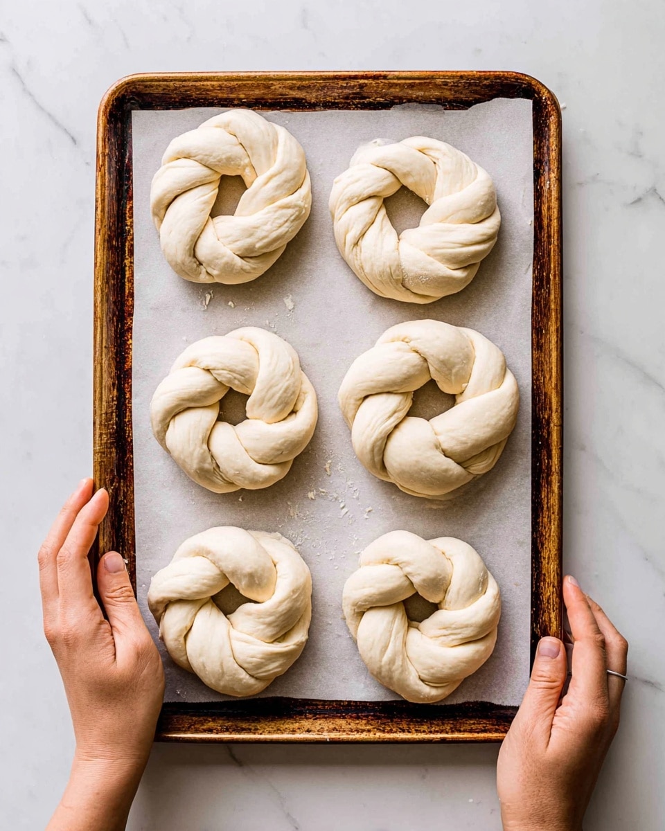 Six uncooked twisted dough rings are placed evenly on a baking tray lined with white parchment paper, each ring made of two thick strands twisted together, showing a smooth, pale dough texture. The tray has a worn, rusty appearance and is held by two woman's hands on a white marbled surface. The lighting is bright and natural, highlighting the soft dough and the rough edges of the tray photo taken with an iphone --ar 4:5 --v 7