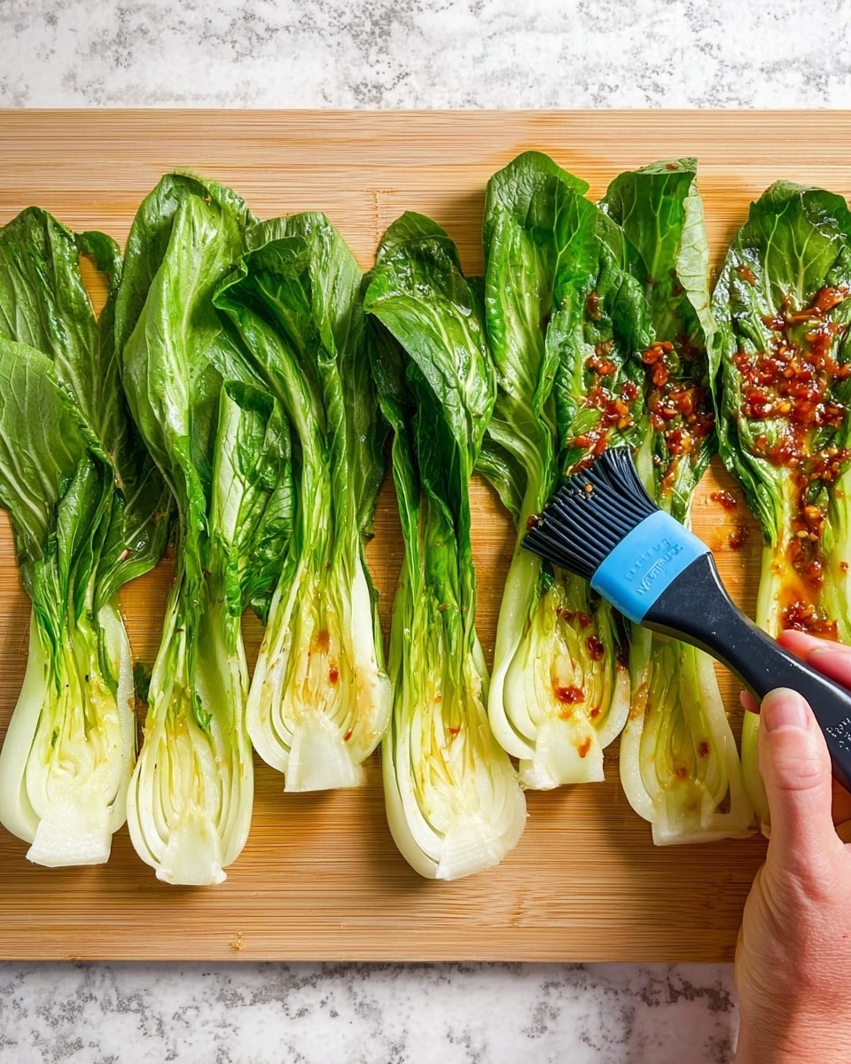 The image shows six pieces of fresh green bok choy arranged in a row on a light wooden cutting board with a white marbled surface underneath. The bok choy is cut in half lengthwise, revealing their white stems and leafy green tops. A woman's hand is holding a black and blue brush, spreading a reddish sauce with small chunks over the white stems and lower leaves of three bok choy halves on the right side. The sauce adds a shiny, glazed look to these pieces, while the three bok choy halves on the left remain plain. photo taken with an iphone --ar 4:5 --v 7