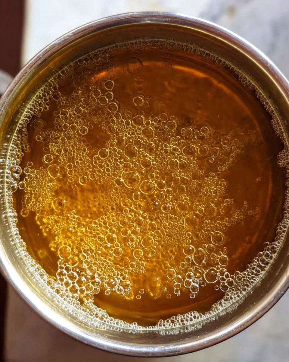 A close-up view inside a silver pot filled with a bubbling golden brown liquid, small round bubbles rising and spreading evenly across the surface, showing a shiny, oily texture under soft lighting, all set on a white marbled surface photo taken with an iphone --ar 4:5 --v 7