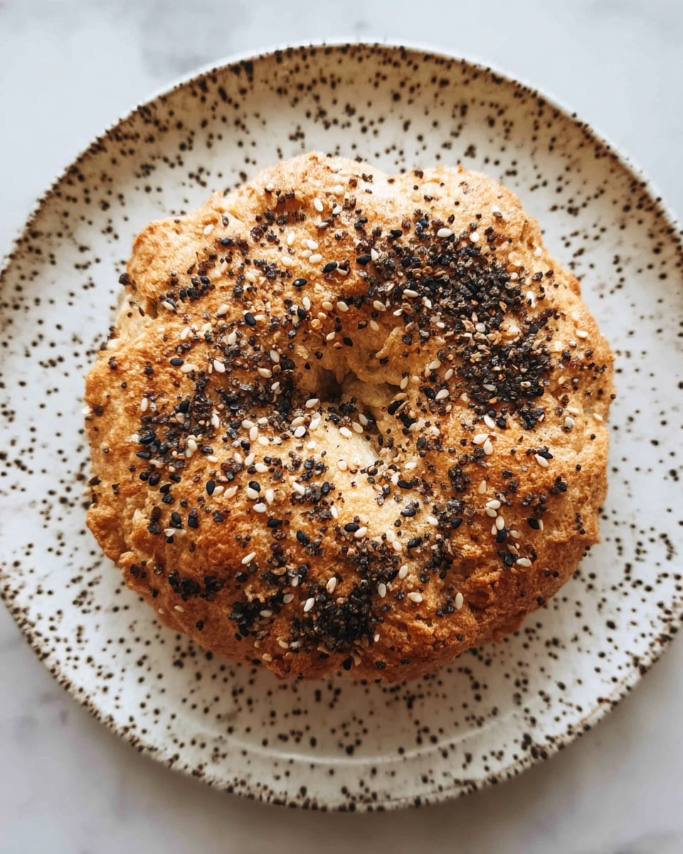 A single round bagel with a bumpy, golden brown crust sits in the center of a white plate with dark speckles. The bagel is topped all over with a mix of black and white sesame seeds and bits of crushed black pepper, adding texture and contrast to its surface. The background is a white marbled texture. photo taken with an iphone --ar 4:5 --v 7