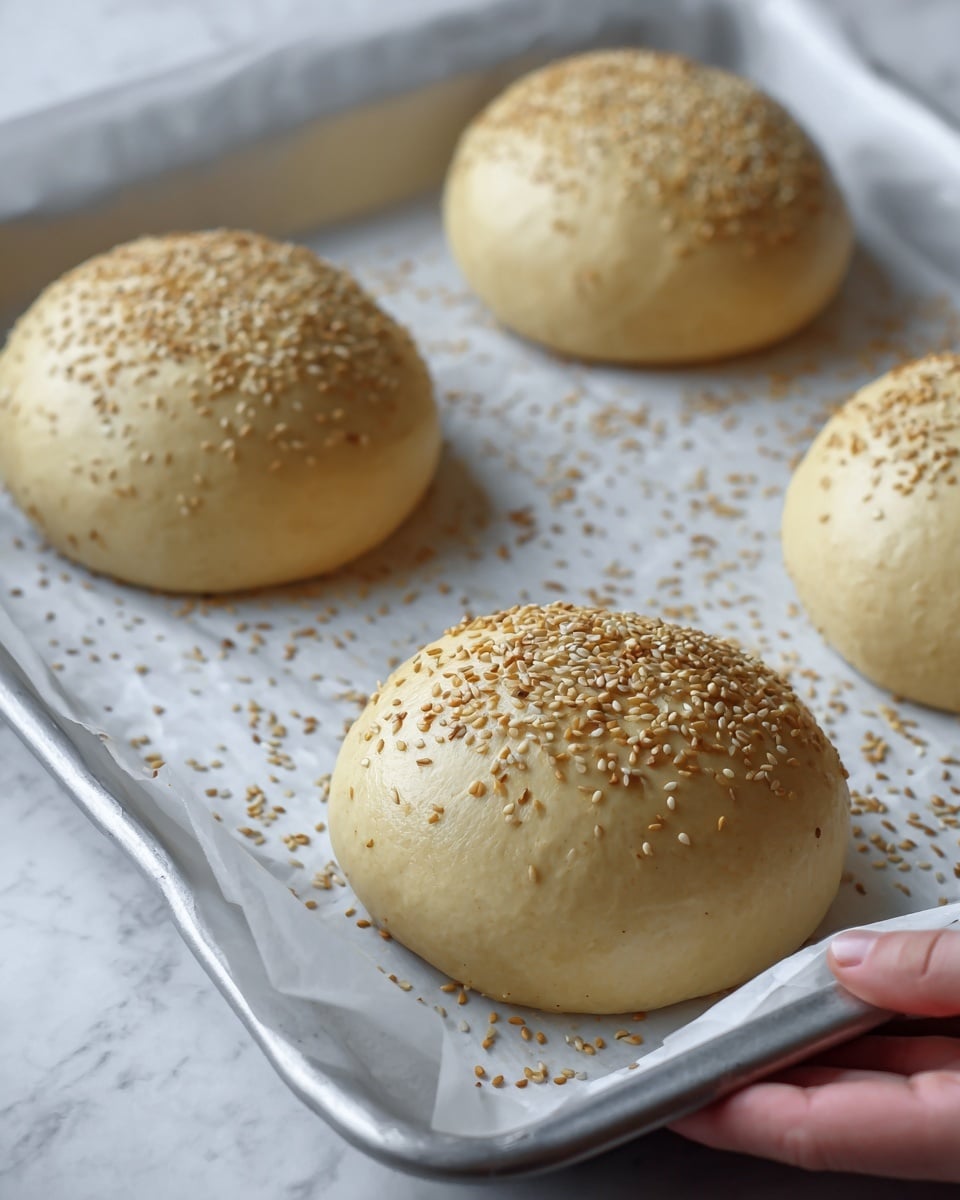 The image shows four round buns on a baking tray lined with white parchment paper, held by a woman's hand. Each bun has a smooth, shiny, light beige dough surface evenly sprinkled with golden sesame seeds. The buns are slightly puffy and spaced apart on the tray. Some sesame seeds are scattered on the parchment paper around the buns. The baking tray has a silver metallic edge, and the background is a white marbled texture. photo taken with an iphone --ar 4:5 --v 7