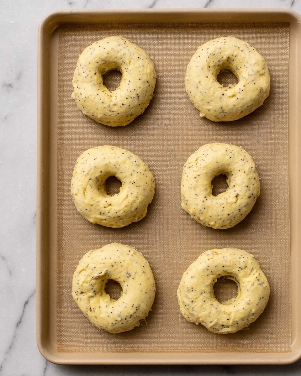 The image shows six dough shapes arranged on a light brown baking sheet with textured paper on a white marbled surface. Each dough piece is round with a visible hole in the center, resembling bagels or doughnuts. The dough has a pale yellow color with small dark specks, possibly seeds or spices, distributed evenly throughout. The texture looks soft and slightly rough. The dough pieces are spaced out neatly, forming two columns and three rows. Photo taken with an iphone --ar 4:5 --v 7