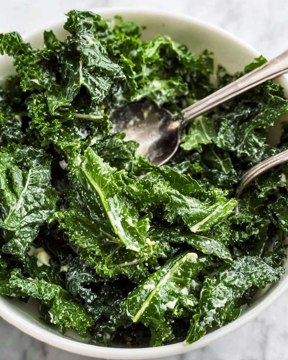 A close-up image of a bowl filled with fresh green kale leaves that are lightly coated with a creamy dressing. The kale leaves have a curly texture and vibrant dark green color, covering most of the bowl with some parts shiny from the dressing. There are two old silver spoons placed inside the bowl among the kale, one partially lifted, showing the layered leaves underneath. The bowl is white and sits on a white marbled surface. photo taken with an iphone --ar 4:5 --v 7