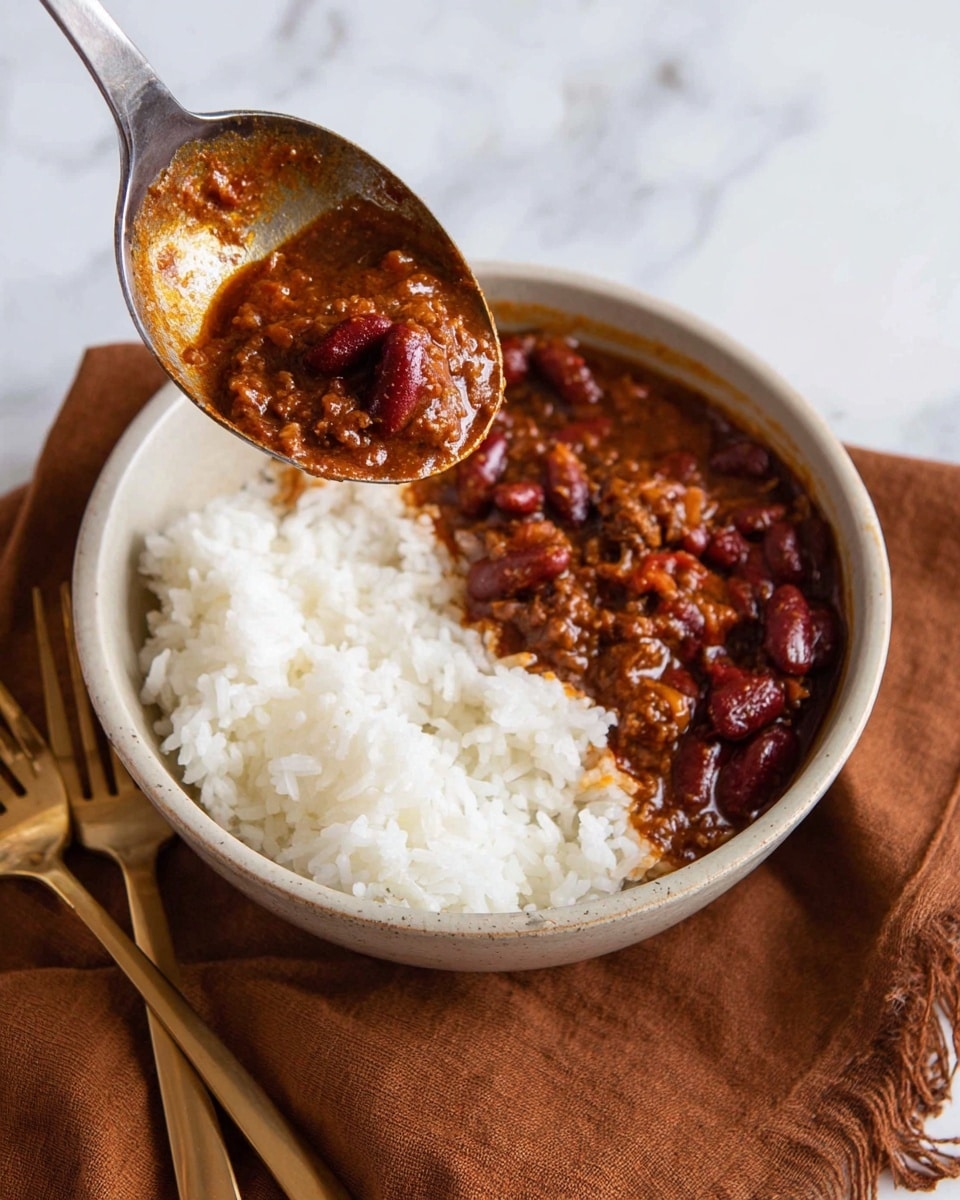 A close-up image showing a white bowl filled halfway with white rice on the left side and thick chili with red kidney beans on the right side. The chili is dark red-brown with chunks of meat and beans, being ladled into the bowl from a large metal spoon held above it. The bowl rests on a brown cloth with a gold fork and spoon beside it on a white marbled surface. Photo taken with an iphone --ar 4:5 --v 7