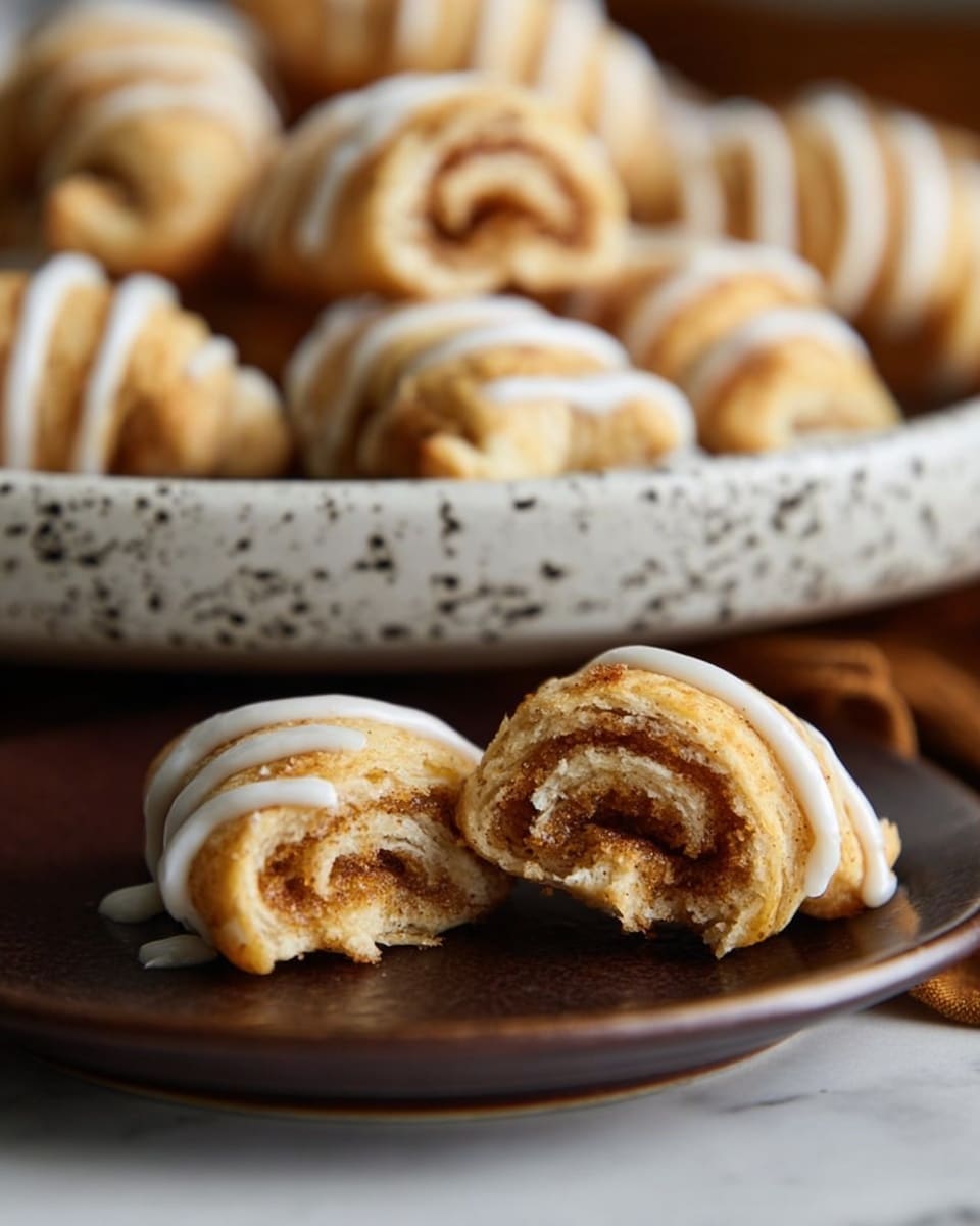The image shows small crescent-shaped pastries with a light golden brown crust and a slightly flaky texture. They have a swirl pattern inside, showing layers of dough and a cinnamon filling. Thin lines of white icing are drizzled over the pastries. One piece is placed in front on a dark brown surface, with the rest on a white speckled plate in the background. The background is a white marbled texture. photo taken with an iphone --ar 4:5 --v 7