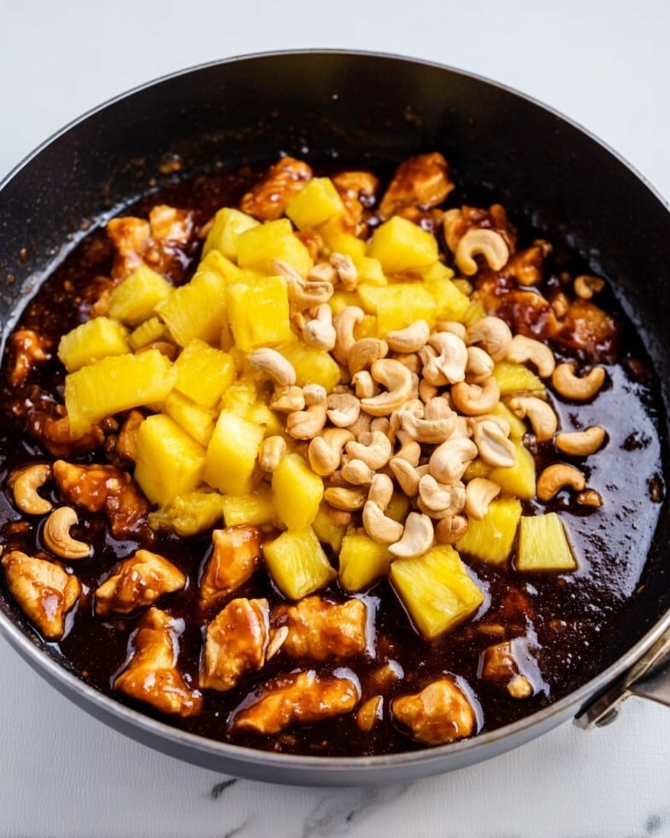 A black pan filled with a dark brown sauce covering small pieces of cooked chicken on the bottom and sides, topped with a pile of bright yellow pineapple chunks in the center, and scattered beige cashew nuts on top of the pineapple. The pan is set on a white marbled surface. photo taken with an iphone --ar 4:5 --v 7