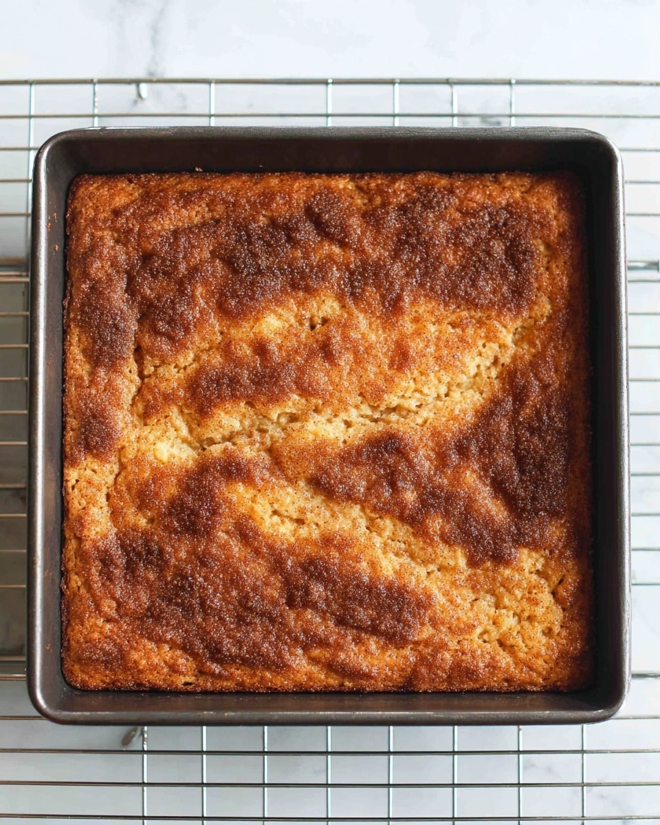 A square baked cake with a rough, golden-brown top layer that has darker cinnamon sugar patches unevenly spread across the surface. The cake is in a dark metal baking pan sitting on a silver cooling rack. The top layer looks slightly cracked and textured with a mix of light and dark brown colors, giving a crispy appearance. The background shows a white marbled texture under the cooling rack. Photo taken with an iphone --ar 4:5 --v 7