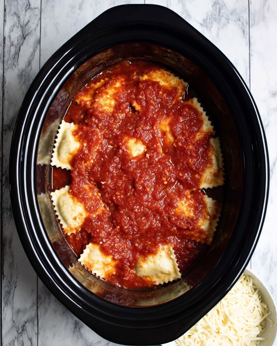 The image shows a black oval slow cooker filled with one layer of square ravioli pasta, covered with a bright red chunky tomato sauce spread evenly on top. The ravioli pieces are pale yellow with ruffled edges and are partially visible through the sauce. The slow cooker is placed on a white marbled surface, and below it, a small white bowl filled with shredded white cheese is partly visible. Photo taken with an iphone --ar 4:5 --v 7