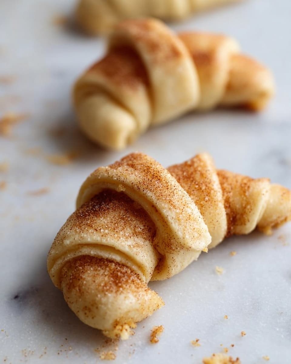 Three raw crescent-shaped dough pieces are placed on a white marbled surface. Each crescent has a light beige color with visible cinnamon sugar sprinkled on and inside the dough, creating a slightly grainy texture. The layers of dough are rolled tightly, showing the spiral shape clearly. The closest crescent is slightly out of focus, the middle one is sharp and detailed, and the farthest crescent is softly blurred. Small crumbs and specks of cinnamon sugar are scattered around the pancakes on the white marbled surface. The photo taken with an iphone --ar 4:5 --v 7