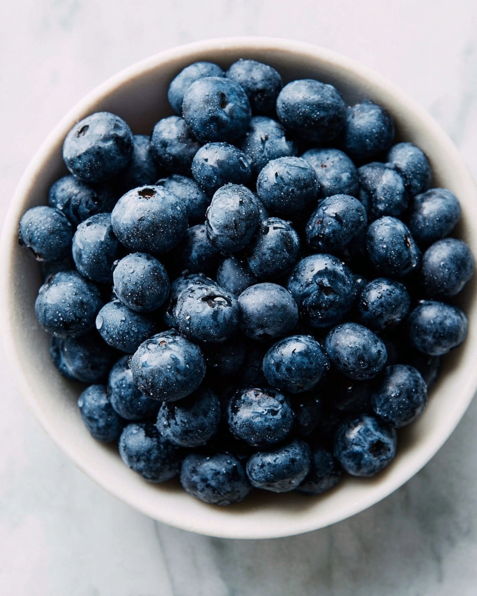 A single layer of fresh blueberries fills a round white bowl placed on a white marbled surface. The blueberries are deep blue with a soft, slightly matte texture, some showing small water droplets that make them look fresh and juicy. The bowl is full and the berries are piled up close to the edge, giving a sense of abundance and freshness. photo taken with an iphone --ar 4:5 --v 7