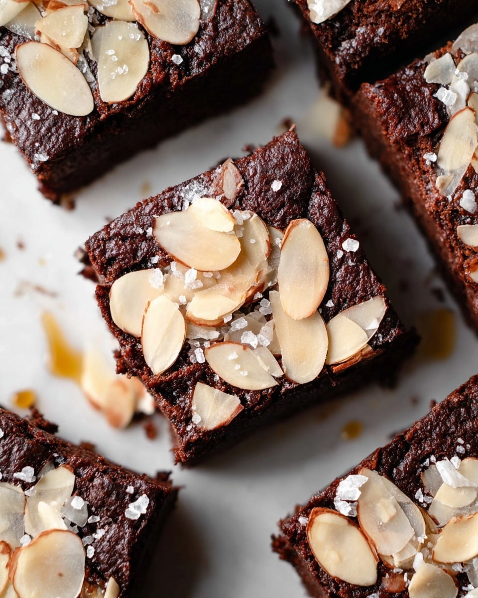 The image shows close-up square brownies with a dark, rich chocolate color and slightly cracked texture on top. Each brownie is topped with thin, pale beige slices of almonds scattered unevenly, giving a crunchy look. Some shiny salt flakes are sprinkled lightly over the nuts, adding a touch of sparkle. The brownies are arranged close to each other on a white marbled surface, with small drips of golden syrup on the side. photo taken with an iphone --ar 4:5 --v 7