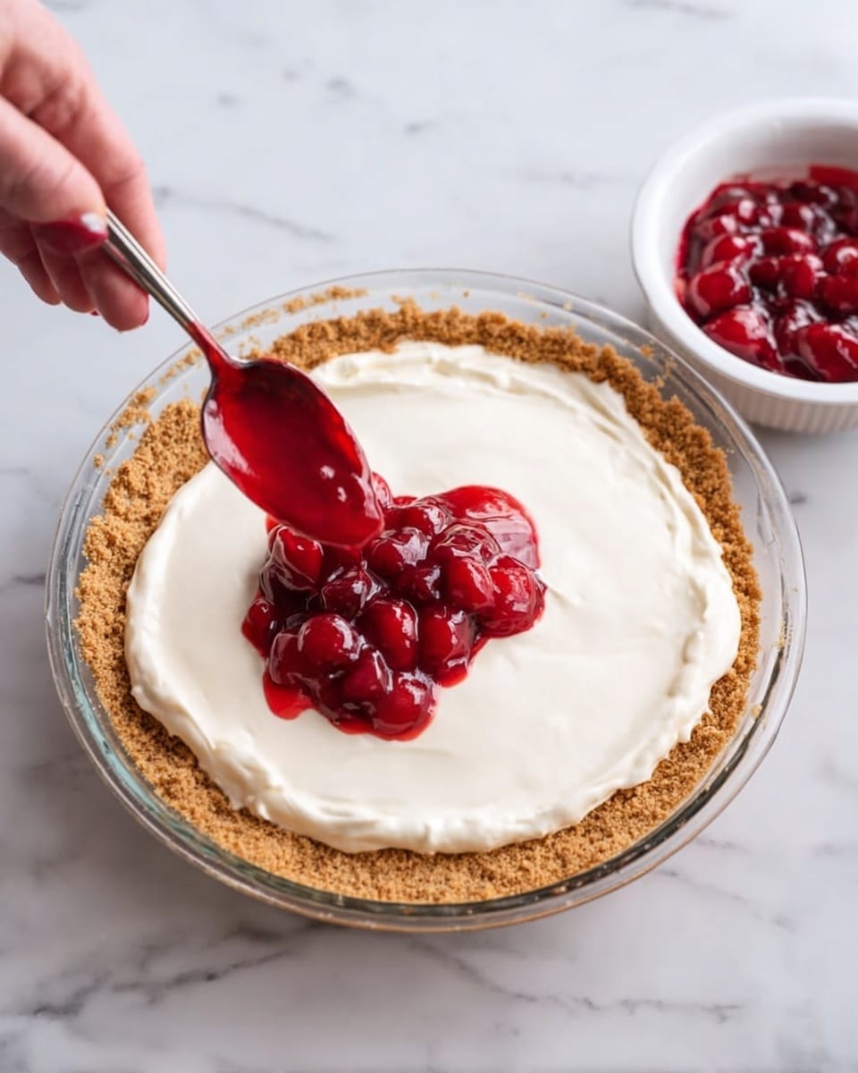A clear glass pie dish holds a dessert with three visible layers: a thick, crumbly light brown crust forming the base and sides, a smooth, creamy white filling spread evenly across the crust, and a shiny red cherry topping being added in the center with a spoon held by a woman's hand above the pie. To the right of the pie, there is a white bowl filled with more of the bright red cherry topping. The whole scene is set on a white marbled surface. photo taken with an iphone --ar 4:5 --v 7