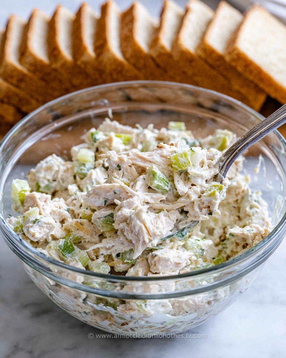 A clear glass bowl contains a creamy chicken salad mixture with visible pieces of shredded white chicken, small chunks of light green celery, and specks of finely chopped herbs, all coated in a thick white mayonnaise-like dressing. A silver fork rests inside the bowl, partially submerged in the mixture. Behind the bowl, there is a row of neatly sliced light brown sandwich bread arranged upright. The setup is placed on a white marbled surface. photo taken with an iphone --ar 4:5 --v 7