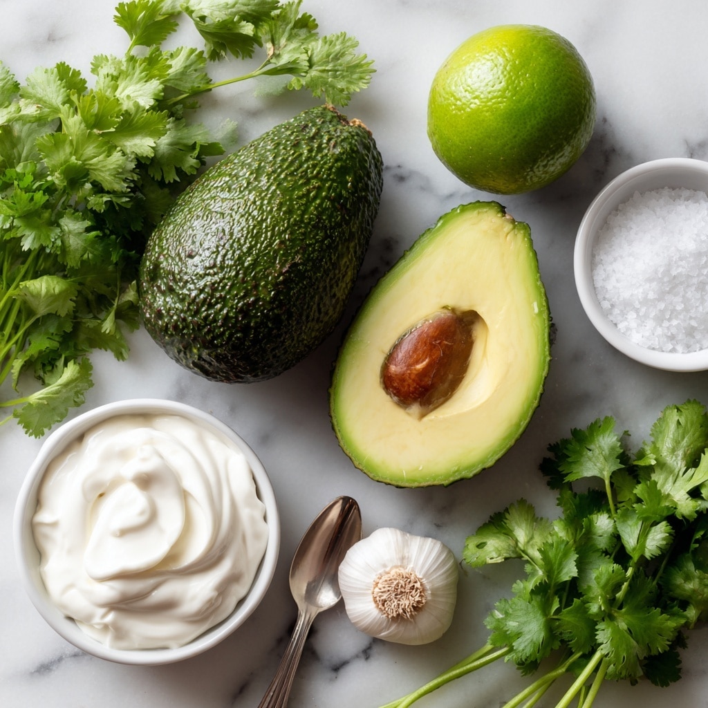 The image shows whole and halved avocados with dark green, bumpy skin and creamy yellow-green flesh, one half containing a large brown seed, placed near a shiny green lime. Below these, there is a silver spoon filled with thick white sour cream on a white marbled surface. Next to it is a small white clove of garlic and a bunch of fresh green cilantro leaves with visible stems. To the right, a small white bowl holds some white salt granules. The items are arranged neatly on the white marbled background. photo taken with an iphone --ar 4:5 --v 7