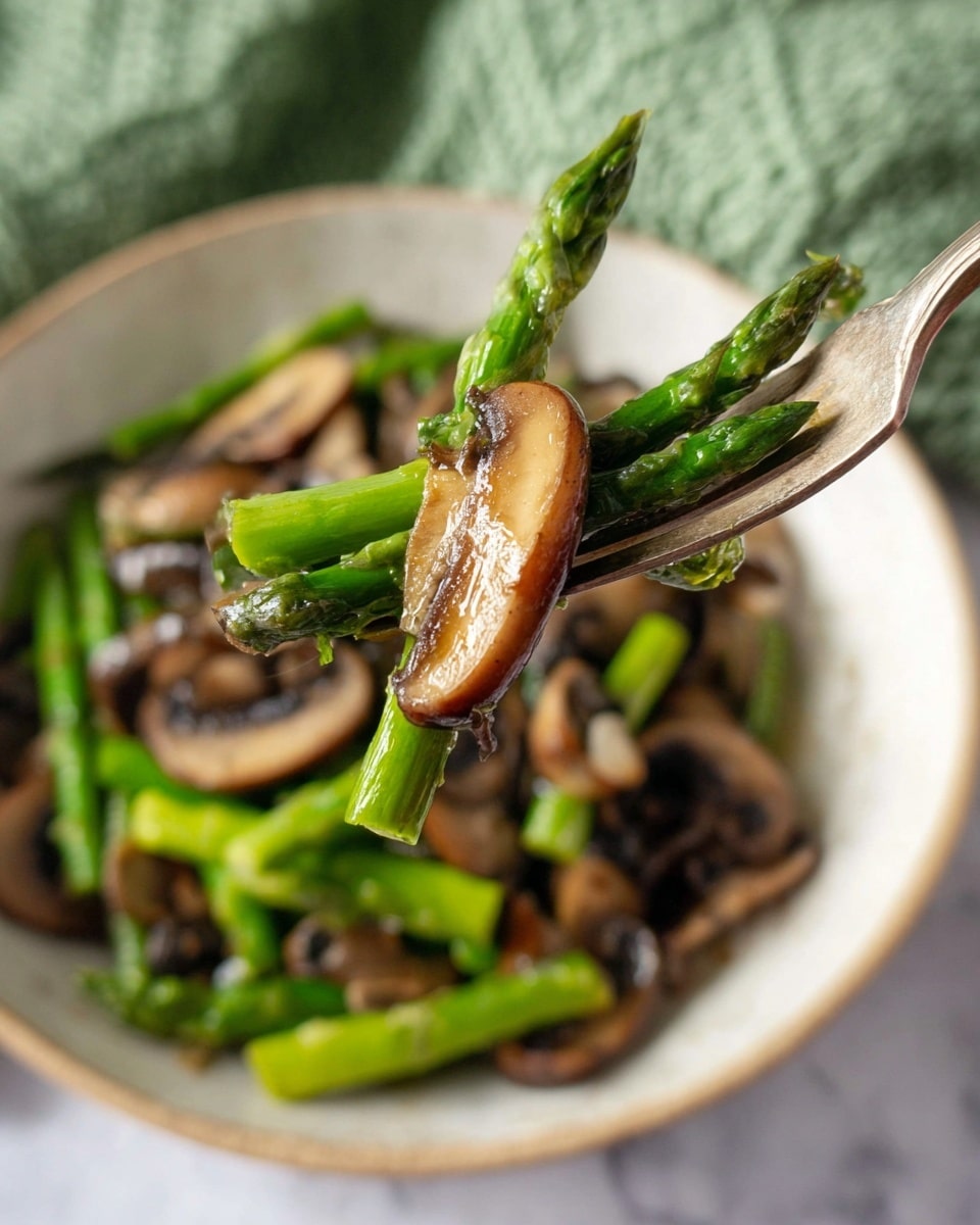 A close-up view shows a fork holding a single bright green asparagus spear and a thin slice of cooked brown mushroom, both shiny with a light glaze. Below the fork, a white bowl is filled with more asparagus pieces and mushrooms, all cooked and mixed together with a glossy texture. The background is a white marbled surface with a soft green knitted cloth partially visible in the top corner. The image shows fresh vegetables with a simple, healthy look. Photo taken with an iphone --ar 4:5 --v 7