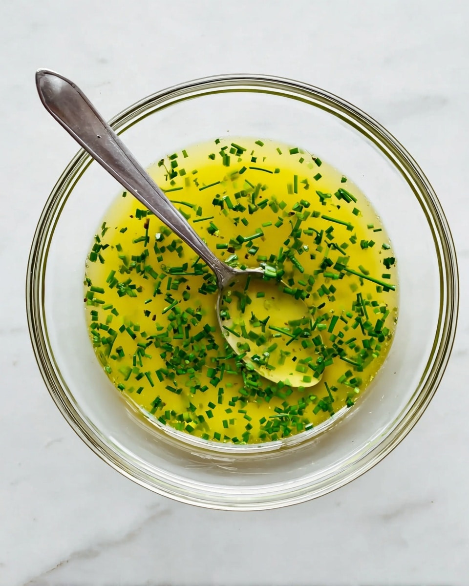 A clear round glass bowl sits on a white marbled surface, filled with a yellow herb dressing scattered with small, finely chopped green chives evenly spread throughout. A shiny silver spoon rests inside the bowl, positioned diagonally from bottom left to top right, partially submerged in the dressing showing the glossy texture of the liquid. The colors are bright with the yellow of the dressing contrasting against the deep green chives, all highlighted by soft natural lighting. Photo taken with an iphone --ar 4:5 --v 7