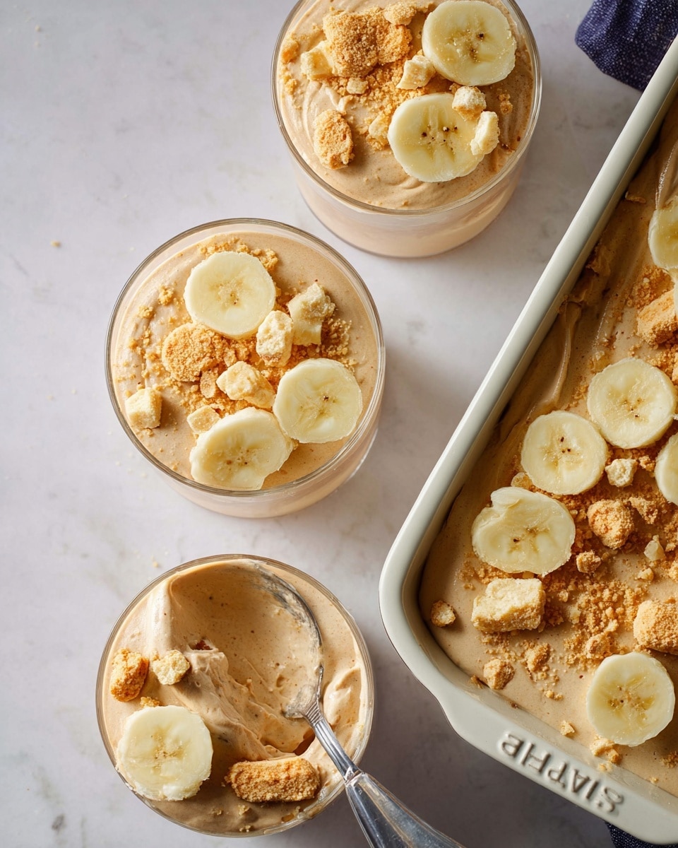 The image shows three clear round glass cups filled with a creamy light brown mousse-like dessert. Each cup is topped with three fresh banana slices and sprinkled with small broken pieces of light yellow crumbly cookies. To the right, there is a white Staub baking dish filled with the same light brown dessert, garnished with banana slices and cookie crumbs. The scene is set on a white marbled surface, and one of the cups in the bottom left corner has a spoon scooping into the dessert, showing the smooth texture inside. Photo taken with an iphone --ar 4:5 --v 7