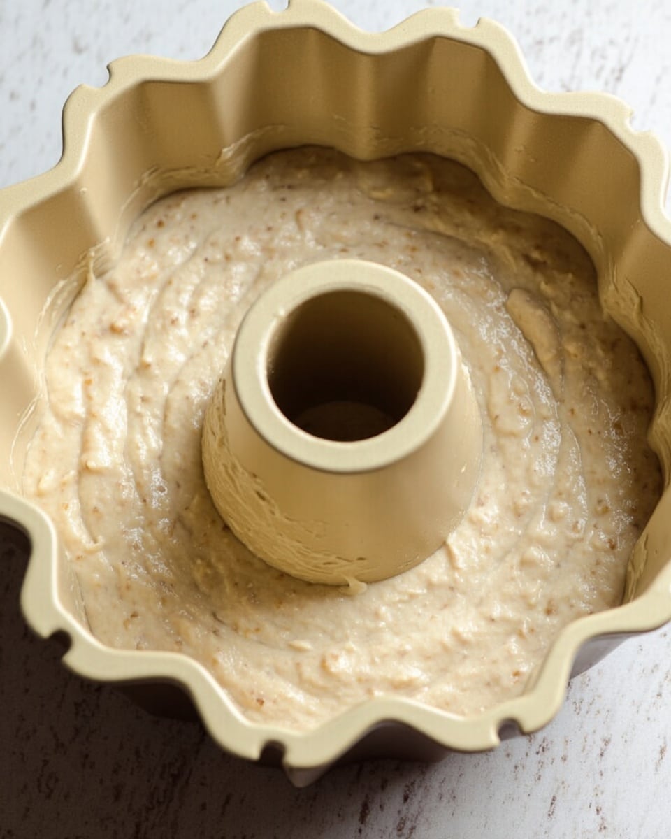 The image shows a light beige batter with small lumps spread inside a cream-colored bundt pan with scalloped edges and a hollow center tube. The batter has a thick and slightly bumpy texture and fills the pan evenly. The pan is placed on a white marbled surface, giving a clean and bright backdrop. The photo focuses closely on the texture and shape of the batter inside the pan. photo taken with an iphone --ar 4:5 --v 7