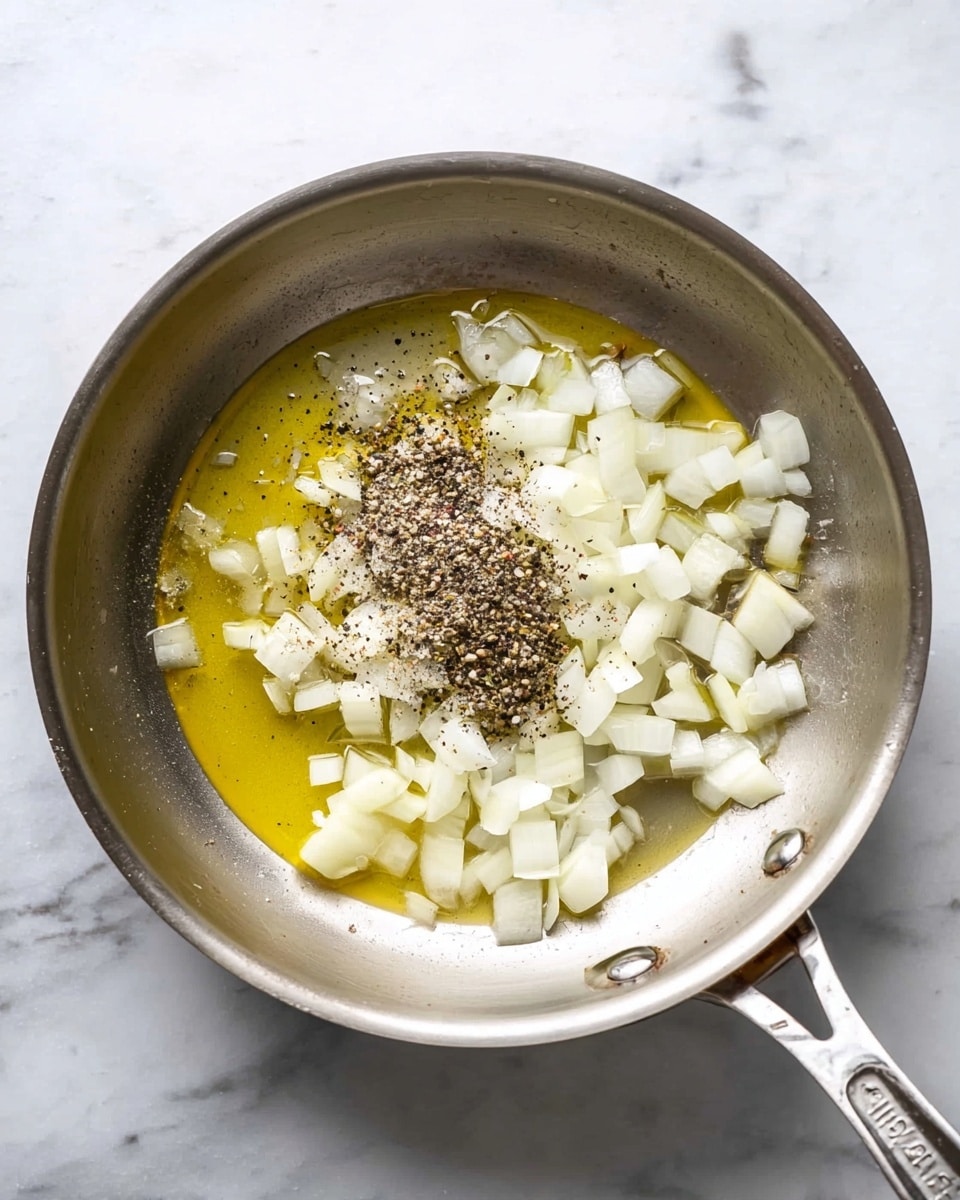 A silver metal pan placed on a white marbled surface contains small white onion cubes spread mostly in the center and right side of the pan. There is a pool of yellow oil under and around the onions, mainly on the left side of the pan. On top of the onion cubes near the center is a small mound of black and white pepper. The pan handle extends out to the left. Photo taken with an iphone --ar 4:5 --v 7
