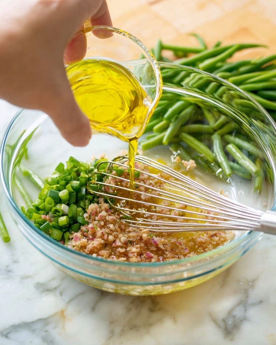A clear glass bowl sits on a white marbled surface with chopped green beans and green onions on the side. Inside the bowl, there is a mixture of light brown minced garlic and shallots, forming the first layer. Over this, a layer of golden olive oil is being poured from a small clear measuring cup held above the bowl. A woman's hand is holding a metal whisk inside the bowl, gently stirring the ingredients. The colors show a mix of translucent brown and bright yellow, with chopped green vegetables in the background adding freshness. Photo taken with an iphone --ar 4:5 --v 7