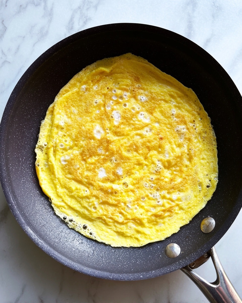 A yellow cooked omelette with small white spots spread thinly and unevenly in a dark non-stick pan, showing a rough, soft texture with some air bubbles near the edges; the pan is placed on a white marbled surface with the handle partly visible at the top right. photo taken with an iphone --ar 4:5 --v 7