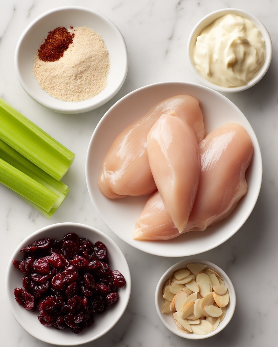 A top view shows several ingredients on a white marbled surface: in the center is a white bowl holding four raw pale pink chicken tenderloins, smooth and slightly shiny. Above and to the right, a white bowl contains creamy, off-white mayo with a soft, thick texture. Above and left of the chicken is a small white bowl with light brown seasoning powder. To the far left, a bright green celery stick lays flat. Below the celery is a small white bowl filled with dark red dried cranberries, round and slightly wrinkled. Below that is another small white bowl with pale slivered almonds, thin and scattered. The scene is neat and bright with clear daylight. Photo taken with an iphone --ar 4:5 --v 7