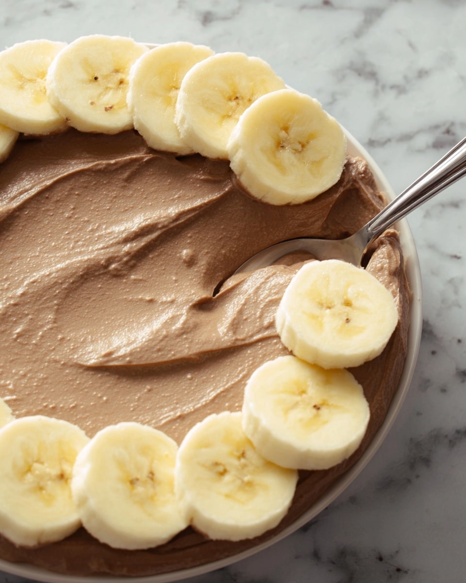 A close-up view of a white plate with a smooth layer of light brown chocolate spread evenly covering the base. On top of the spread, there is a row of sliced bananas, light yellow with soft texture, placed evenly around the edge of the plate. A metal spoon is gently smoothing the chocolate spread near the bottom edge, showing the creamy, thick texture. The background features a white marbled surface. Photo taken with an iphone --ar 4:5 --v 7