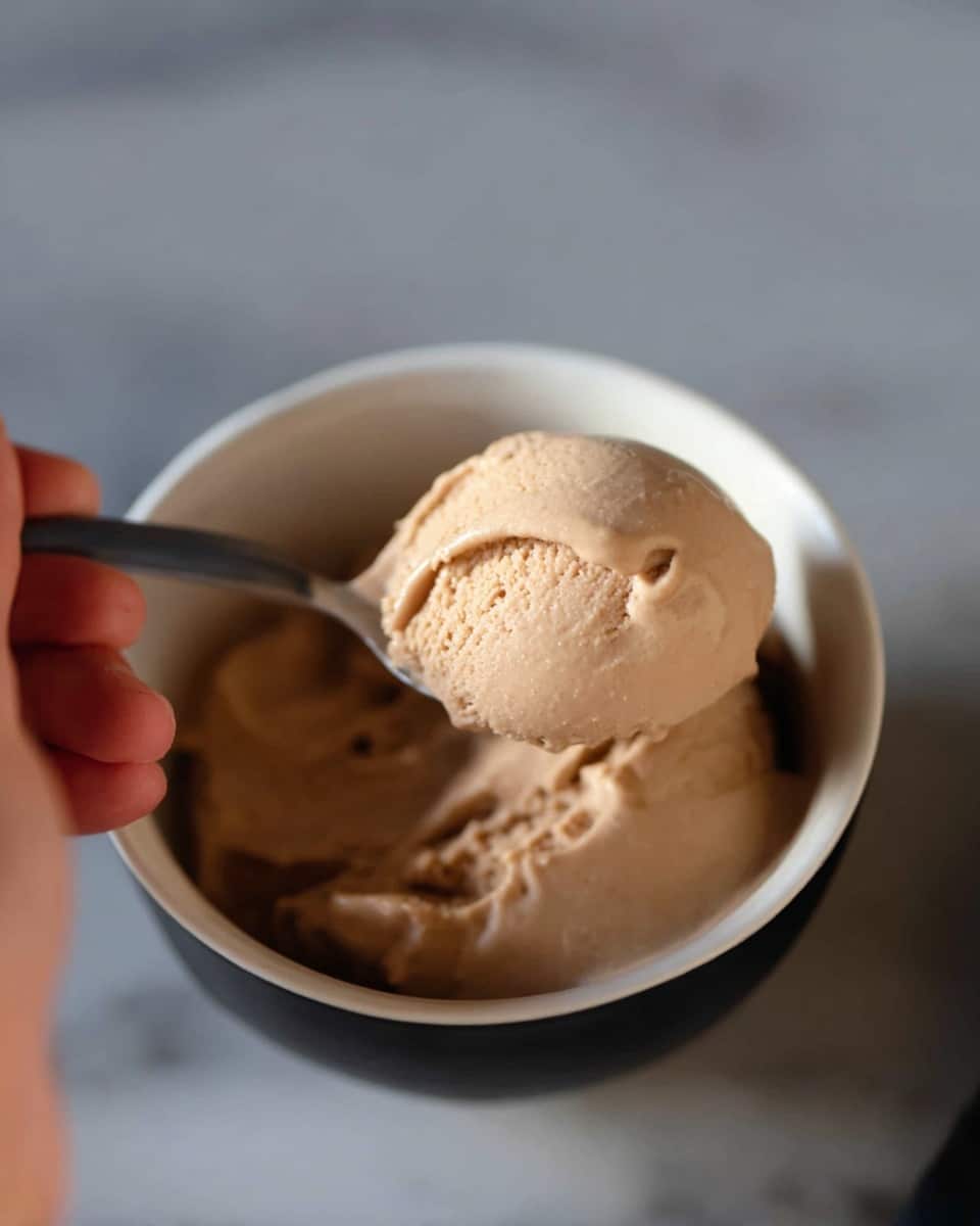 A close-up image of a single scoop of light brown ice cream with a smooth, creamy texture sitting inside a white bowl; a woman's hand is holding a metal spoon scooping some ice cream from the bowl, with a soft blur background and the scene is placed on a white marbled surface. photo taken with an iphone --ar 4:5 --v 7
