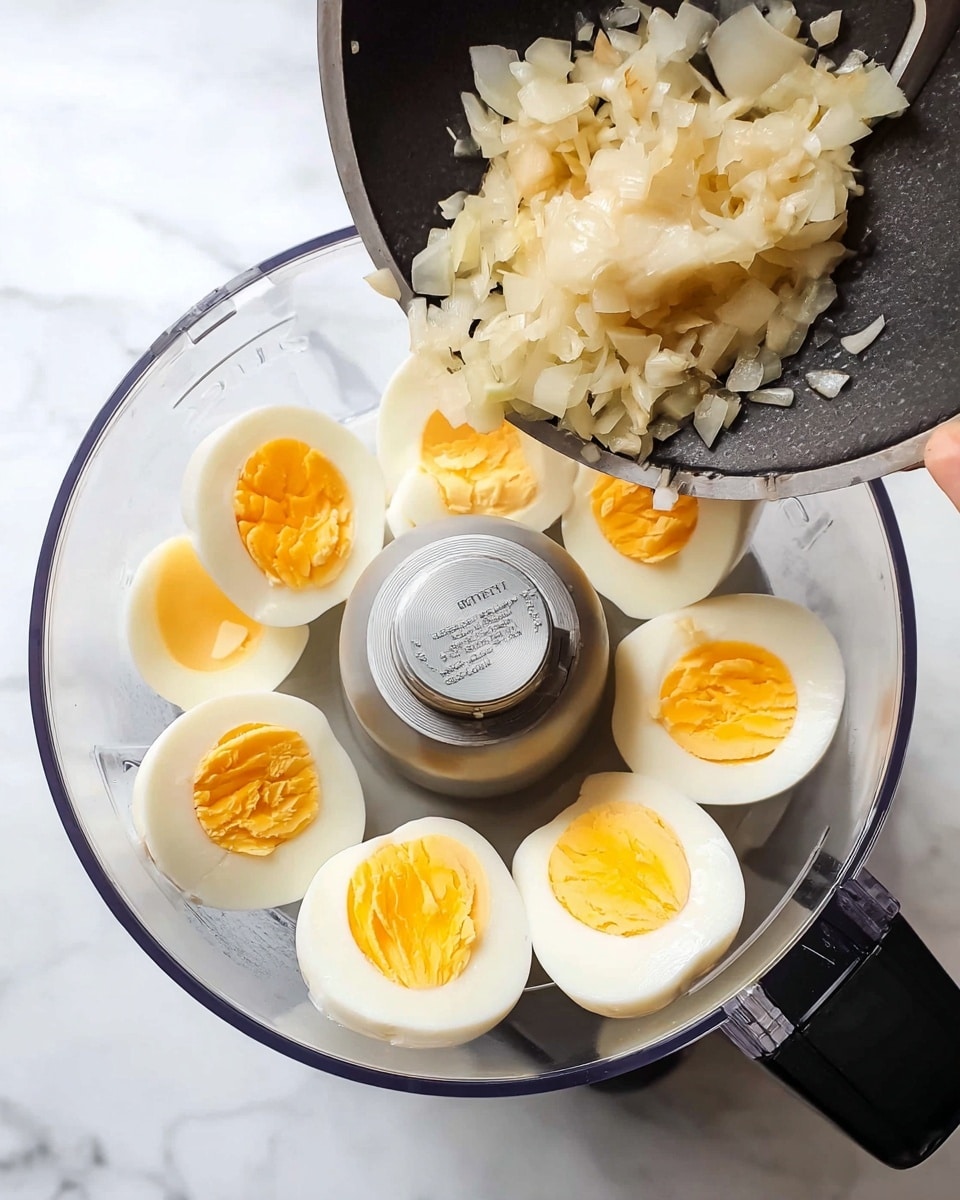 A clear food processor bowl sits on a white marbled surface, containing six halves of hard boiled eggs with bright yellow yolks and smooth white edges arranged around the central blade. Above the bowl, a woman's hand holds a black pan that is pouring small, pale pieces of cooked onions into the food processor. The scene is bright and clean, showing the raw textures of the eggs and the translucent, slightly soft cooked onions about to be mixed, photo taken with an iphone --ar 4:5 --v 7