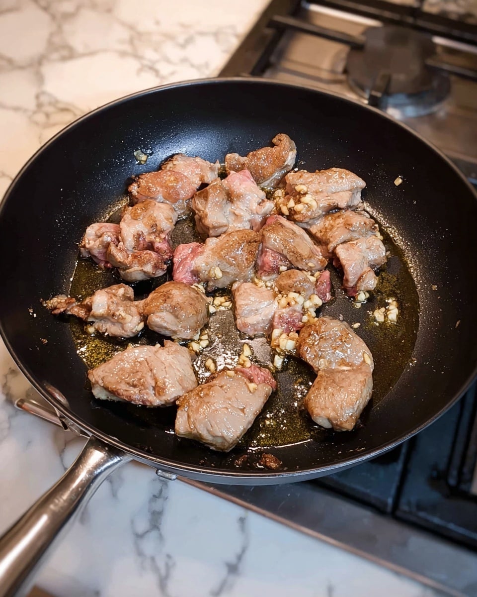 The image shows a black frying pan on a stove with several pieces of meat cooking inside. The meat pieces vary in size and shape, with a light brown color from cooking, and some pink parts still visible. There are small bits of what looks like minced garlic spread around the meat, sizzling in the oil that coats the pan’s surface. The pan handle is silver and extends to the left, and the stove and a white marbled countertop are visible around the pan. Photo taken with an iphone --ar 4:5 --v 7