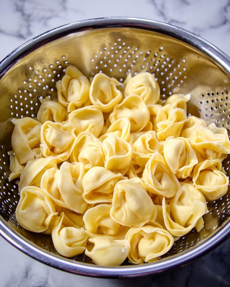 This image shows a metal colander filled with many cooked tortellini pasta pieces. The tortellini are light yellow and smooth, with a slightly shiny surface. They are folded into small ring shapes with pinched edges, filling the colander almost to the top. The colander itself is silver and has many small holes for draining water. The background is a white marbled texture. photo taken with an iphone --ar 4:5 --v 7