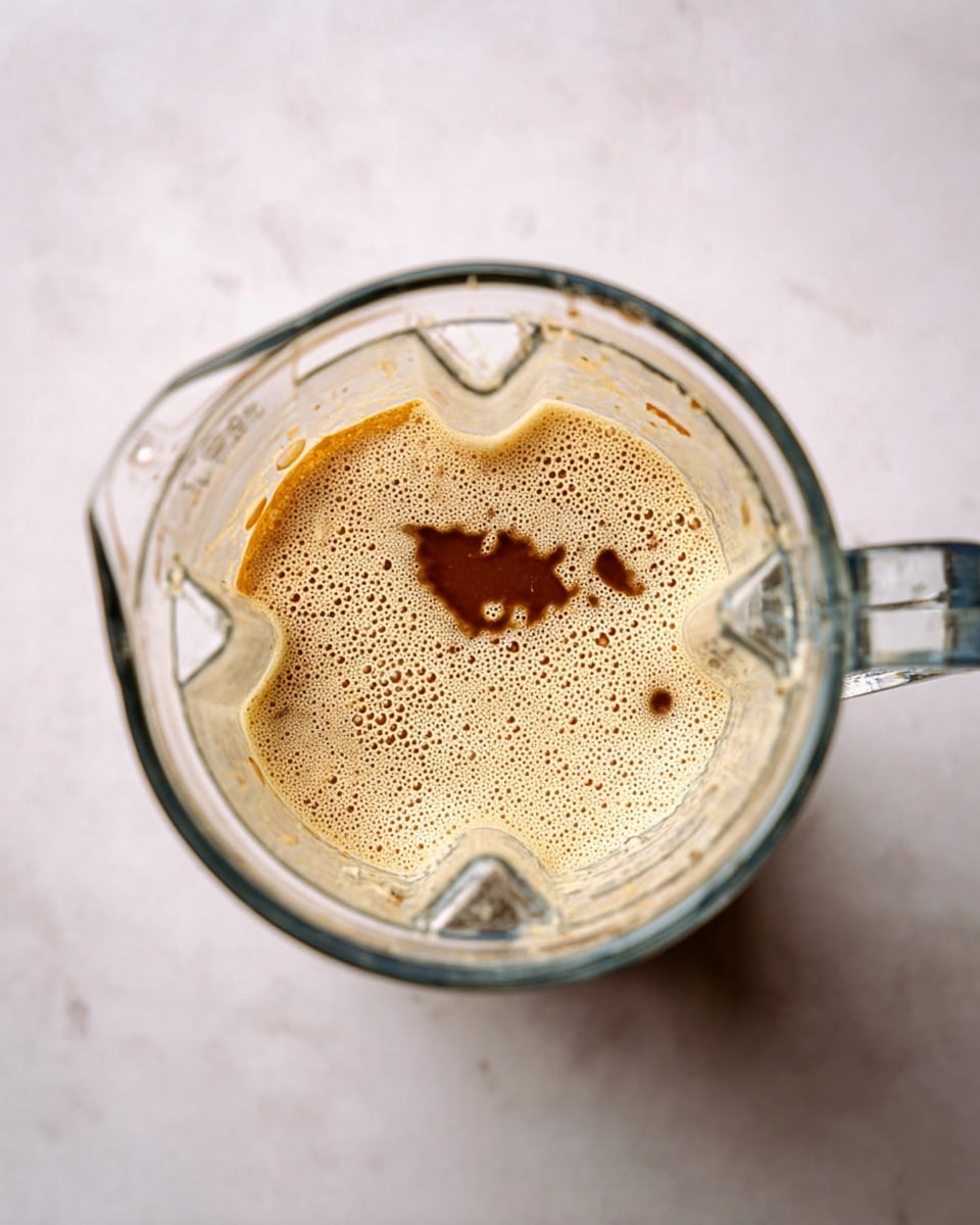 The image shows a clear glass blender jar from above, filled with a foamy light beige liquid layer with bubbles covering most of the surface. There are two darker spots showing slightly mixed brown liquid in irregular small shapes on the foam. The jar is placed on a white marbled surface with subtle light patterns. The blender jar’s handle is visible on the right side. photo taken with an iphone --ar 4:5 --v 7