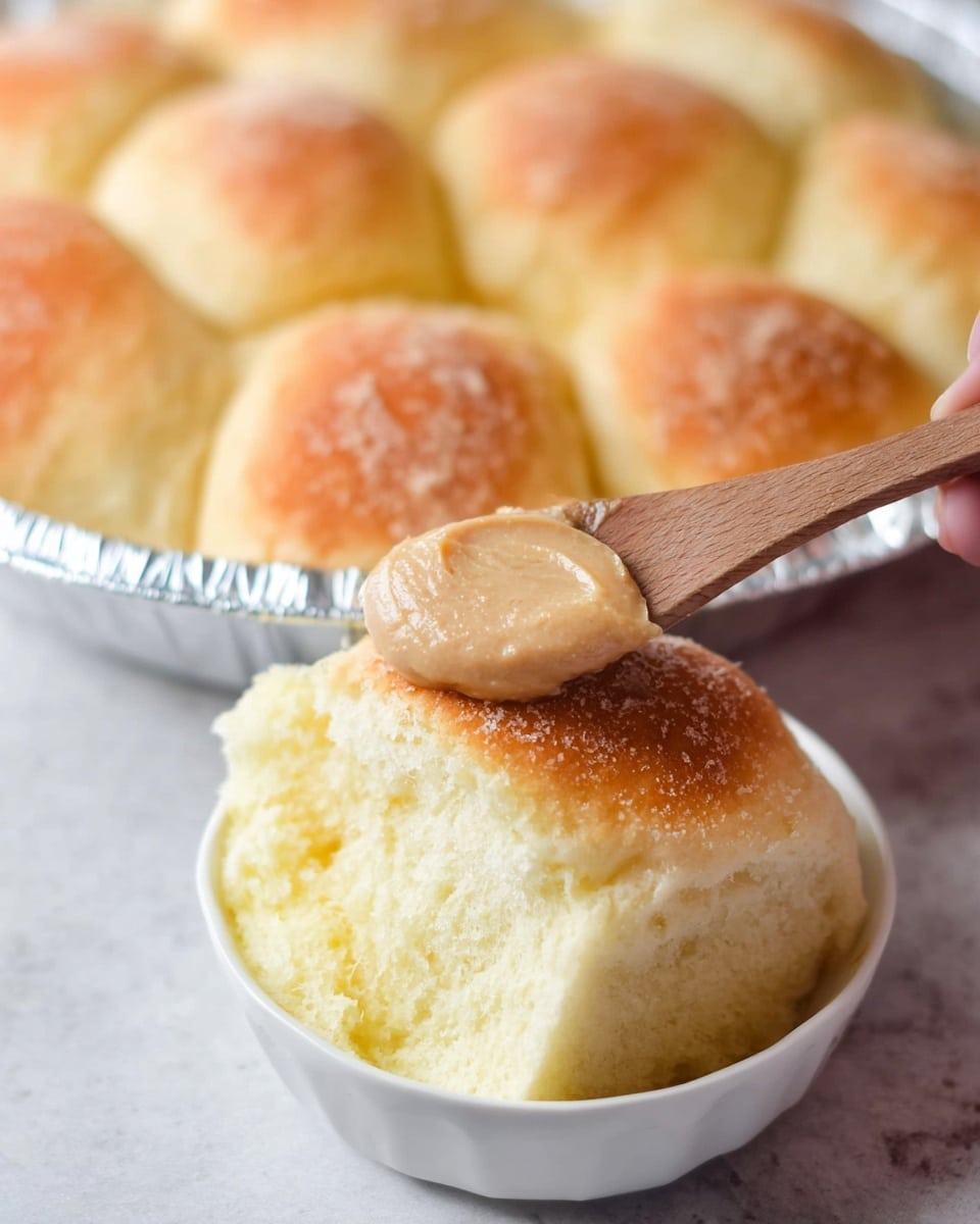 A close-up of a soft yellow bread roll placed in a small white bowl on a white marbled surface, with a smooth light brown spread being scooped onto the bread by a wooden spoon held by a woman's hand. In the background, a round foil tray holds several soft bread rolls with a light golden crust on top, slightly blurred. The bread looks fluffy and airy with a spongy texture. Photo taken with an iphone --ar 4:5 --v 7