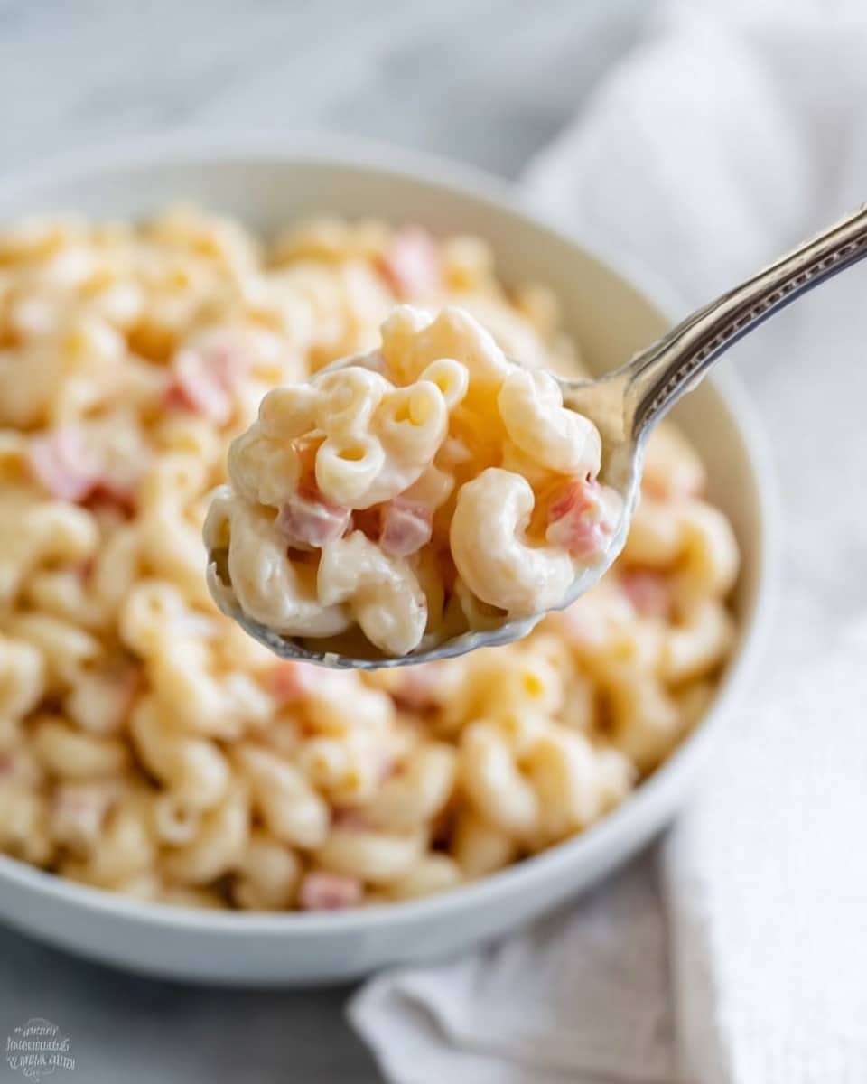 A close-up of a silver spoon holding a few soft, pale yellow macaroni pasta pieces covered in a creamy light-colored sauce with small bits of pink ham mixed in. In the background, a white bowl filled with the same macaroni and ham mixture is slightly out of focus, sitting on a white marbled surface with a white cloth partially visible. The sauce looks smooth and shiny, coating each pasta piece evenly. photo taken with an iphone --ar 4:5 --v 7