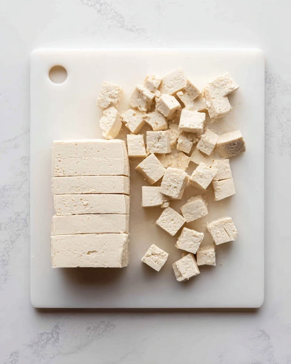 The image shows a white bowl with a black rim filled with beige tofu cubes sprinkled with white powder, likely flour or starch, inside it. A clear spatula with a red silicone head is resting among the tofu cubes inside the bowl. The bowl is placed on a surface with a white marbled texture, which gives a clean and fresh look to the setting. photo taken with an iphone --ar 4:5 --v 7