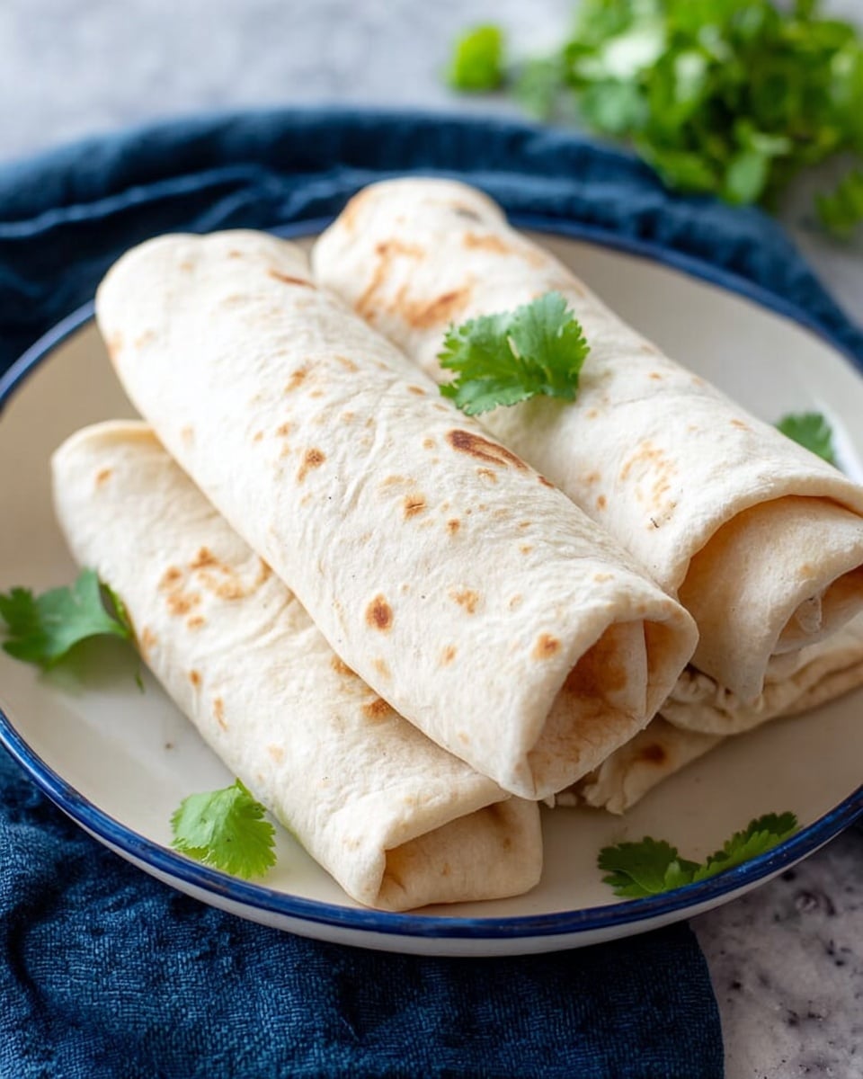 Three rolled tortillas with a light beige color and soft texture are placed neatly on a white plate with blue rim. Each tortilla has small brown spots from cooking. A few green cilantro leaves rest on top of the middle tortilla and are scattered around the plate for decoration. The plate sits on a dark blue cloth with a blurred green garnish in the background against a white marbled surface. The photo taken with an iphone --ar 4:5 --v 7