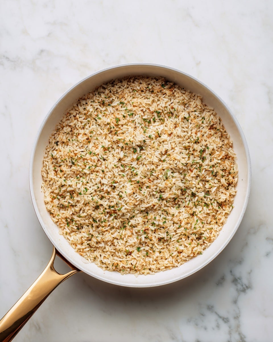 A white pan with a golden handle sits on a white marbled surface, filled evenly with a single layer of cooked brown rice mixed with small bits of herbs and spices, giving it a textured and slightly speckled look. The rice covers the pan’s bottom fully in a flat circle, showing some green and darker brown flecks scattered throughout. The pan is clean with some shadows beneath the handle, creating a simple and neat presentation. photo taken with an iphone --ar 4:5 --v 7