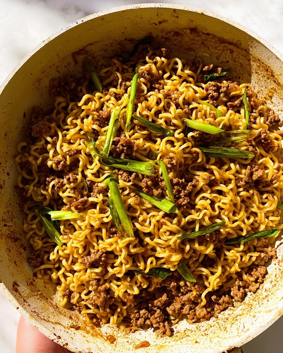 This image shows a close-up of a cooked noodle dish in a light-colored pan with visible sauce stains on the sides. The dish has two main layers: yellow curly noodles and small pieces of cooked ground meat mixed throughout. Scattered on top are thin slices of bright green vegetables, likely green onions, adding a fresh touch. The nails of a woman's hand can be seen holding the pan lightly at the edge. The noodles have a slight shine from the sauce, and the texture appears soft yet firm. The background has a clean white marbled texture. photo taken with an iphone --ar 4:5 --v 7
