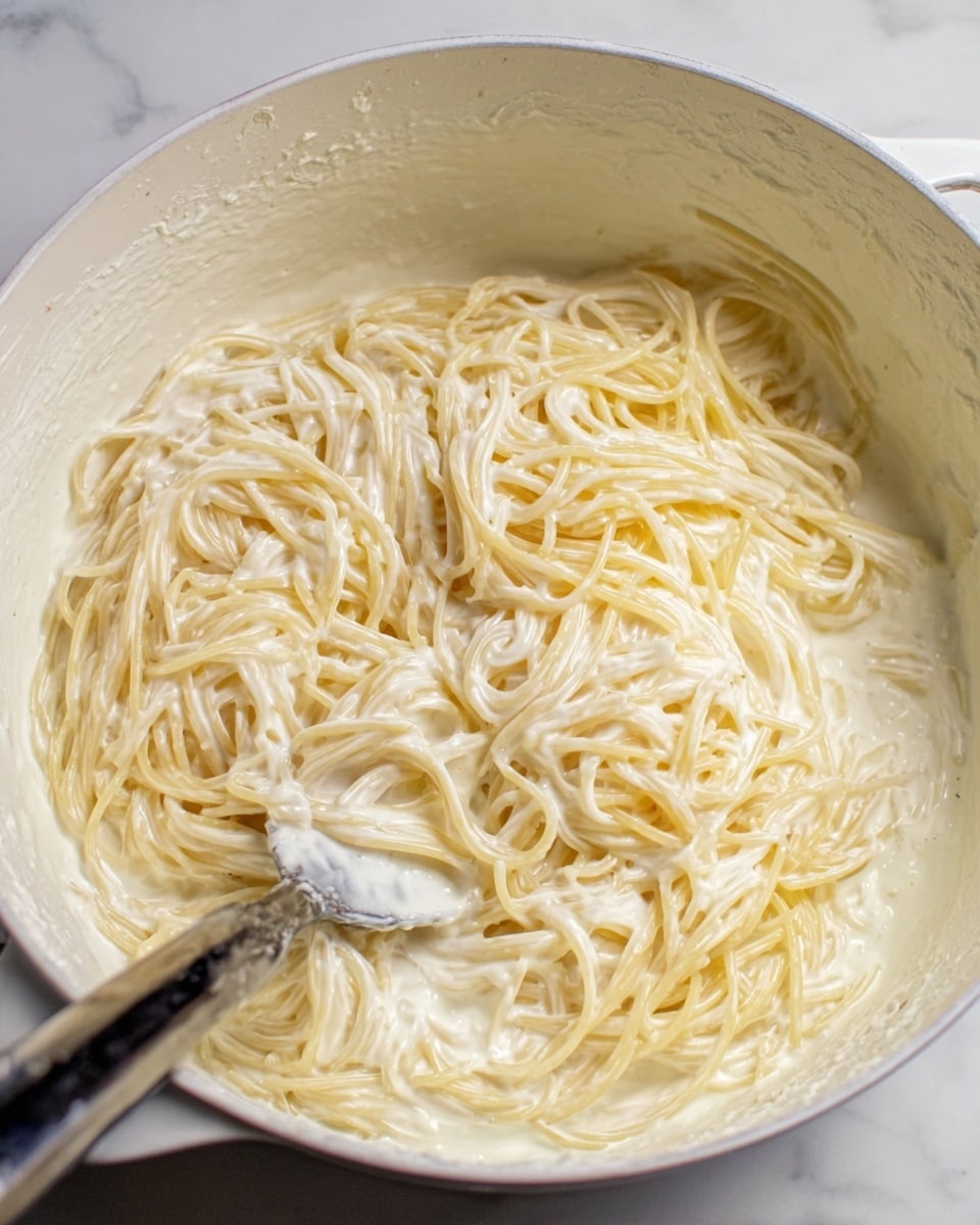 A white pot filled with creamy spaghetti pasta, the noodles coated in a thick, smooth white sauce. The spaghetti strands are loosely piled in the center, with some strands spread out around the sides. A metal spoon with a dark handle is partially visible on the left side, slightly covered in the creamy sauce, resting inside the pot. The inside surface of the pot shows some sauce splatters, blending with the white color of the pot. The photo is taken on a white marbled surface. photo taken with an iphone --ar 4:5 --v 7