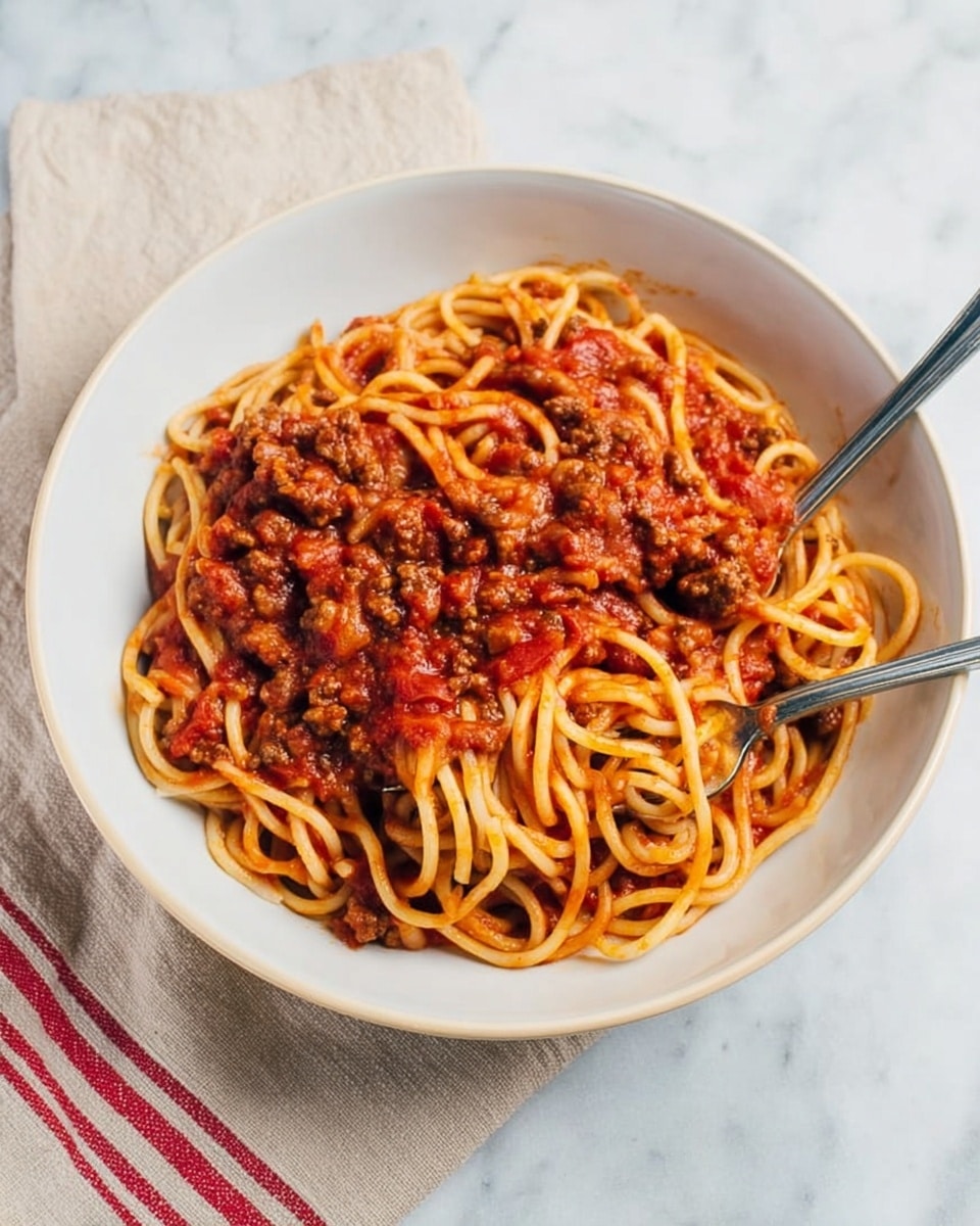 A white bowl filled with thick spaghetti noodles mixed with chunky red tomato sauce and browned ground meat pieces, creating a rich, textured top layer of sauce and meat covering the pasta beneath. Two silver forks rest diagonally on the left side inside the bowl, slightly tangled with the noodles. The bowl is placed on a white marbled surface with a beige cloth napkin edged with red stripes to the left. Photo taken with an iphone --ar 4:5 --v 7