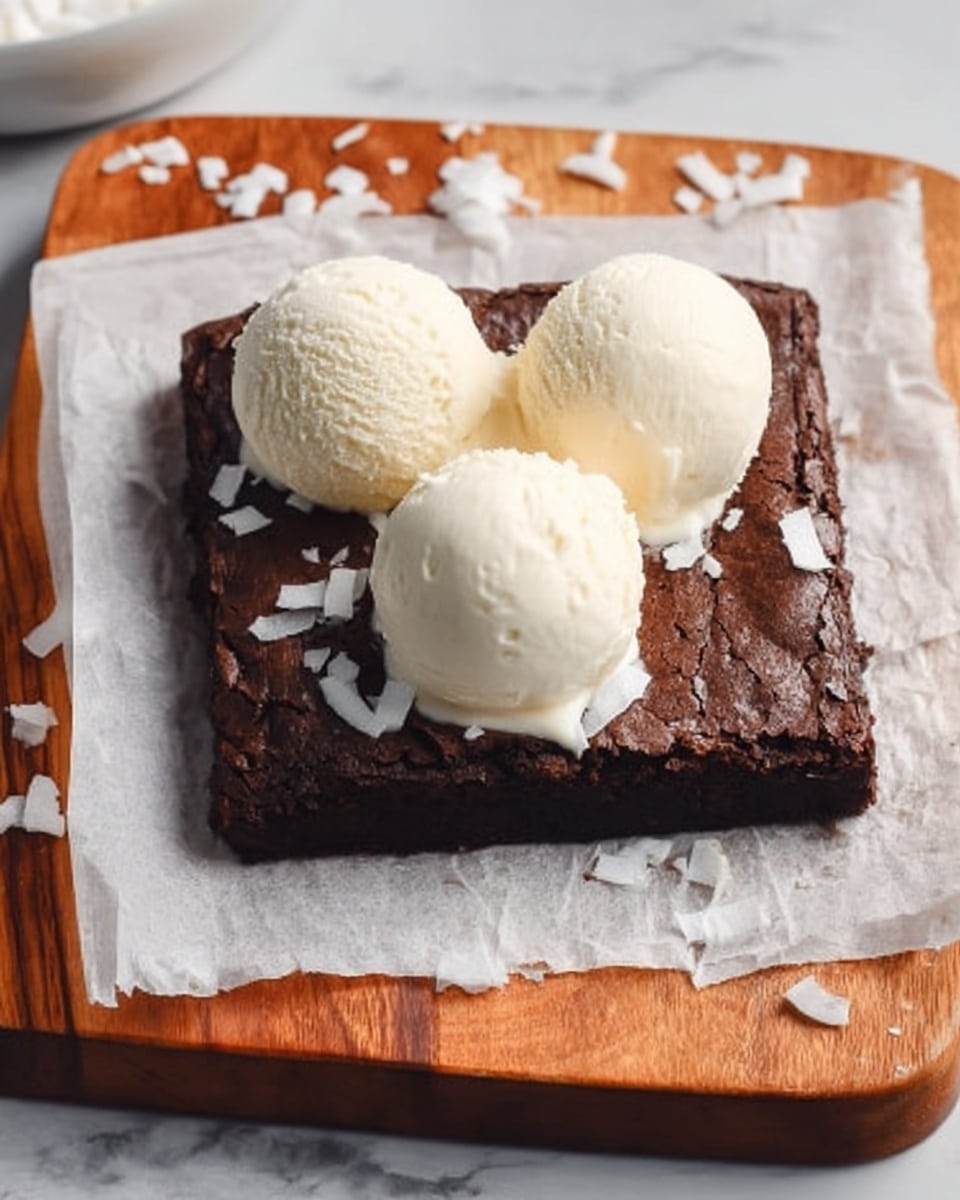 A square dark brown brownie with a cracked top sits on a white marbled surface covered with parchment paper on a wooden cutting board. There are three smooth, round scoops of pale cream-colored ice cream placed evenly on top of the brownie. Around the brownie, there are scattered white coconut flakes for decoration. photo taken with an iphone --ar 4:5 --v 7