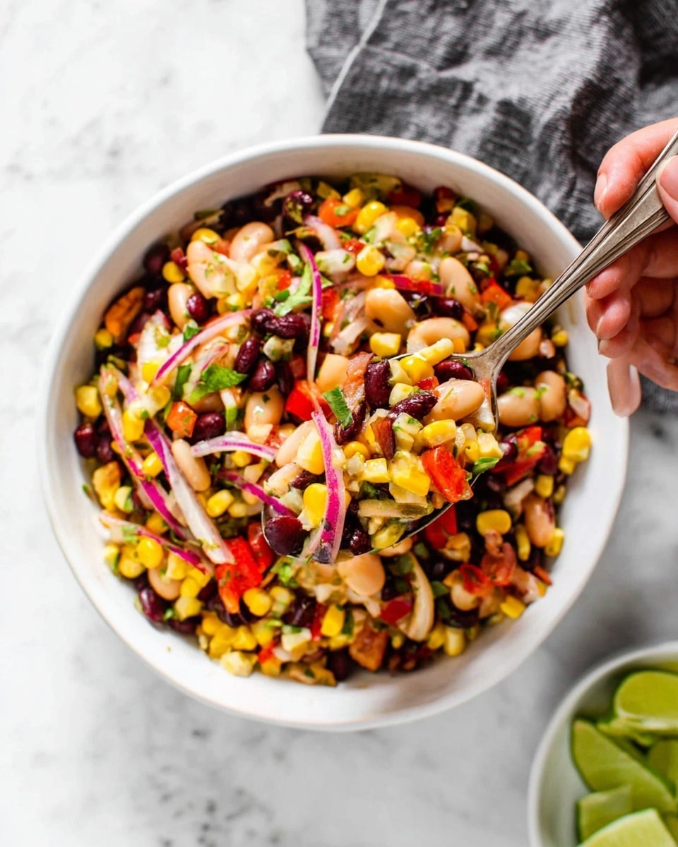 A white bowl filled with a colorful mixed bean salad showing many layers of color and texture. The mixture contains dark brown black beans, white beans, yellow corn kernels, thin red onion slices, green herbs, and small pieces of red bell pepper. A woman's hand holds a spoon full of this mixture, lifting it up from the bowl. The background has a white marbled surface with a gray cloth nearby, and a small white dish in the corner holds green lime wedges. The image gives a fresh, vibrant, and healthy feeling. photo taken with an iphone --ar 4:5 --v 7
