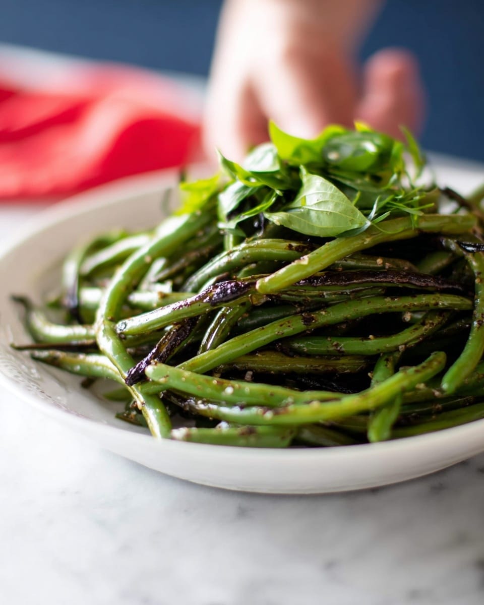 A white plate holds a pile of cooked green beans that have a slight char with darker brown spots. The beans vary in length and are mixed with bright green fresh leaves scattered on top and nestled among them. The beans have a shiny, slightly oily texture, and the plate is placed on a white marbled surface with a blurred background showing a lady's hand and red color. photo taken with an iphone --ar 4:5 --v 7