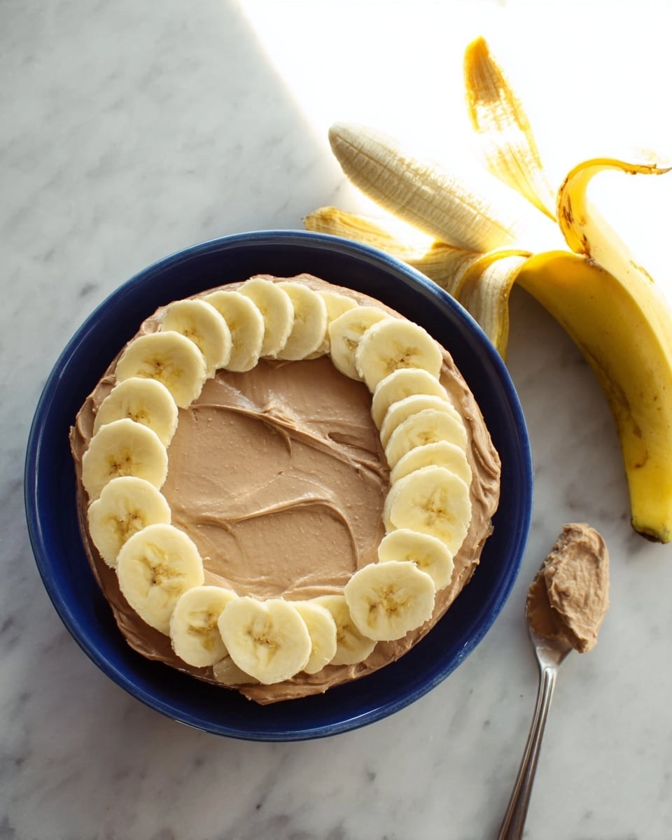 A deep blue round bowl filled with one visible layer of smooth, creamy light brown spread, evenly covering the base, topped with a ring of evenly spaced, thinly sliced banana pieces that are light yellow with a soft texture, all placed on a white marbled surface. To the right, there is a peeled banana resting on a banana peel, with a metal spoon holding some of the light brown spread resting on the peel. photo taken with an iphone --ar 4:5 --v 7