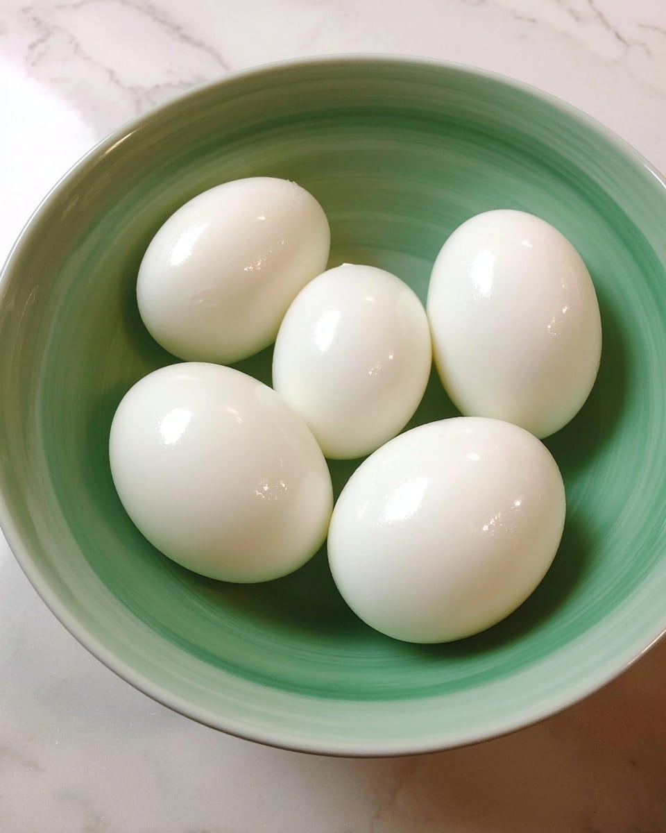 Five smooth white hard-boiled eggs are placed inside a white bowl with a soft green inner color. The eggs have a shiny surface reflecting light evenly. The bowl sits on a white marbled surface, adding a clean and simple look to the scene. photo taken with an iphone --ar 4:5 --v 7