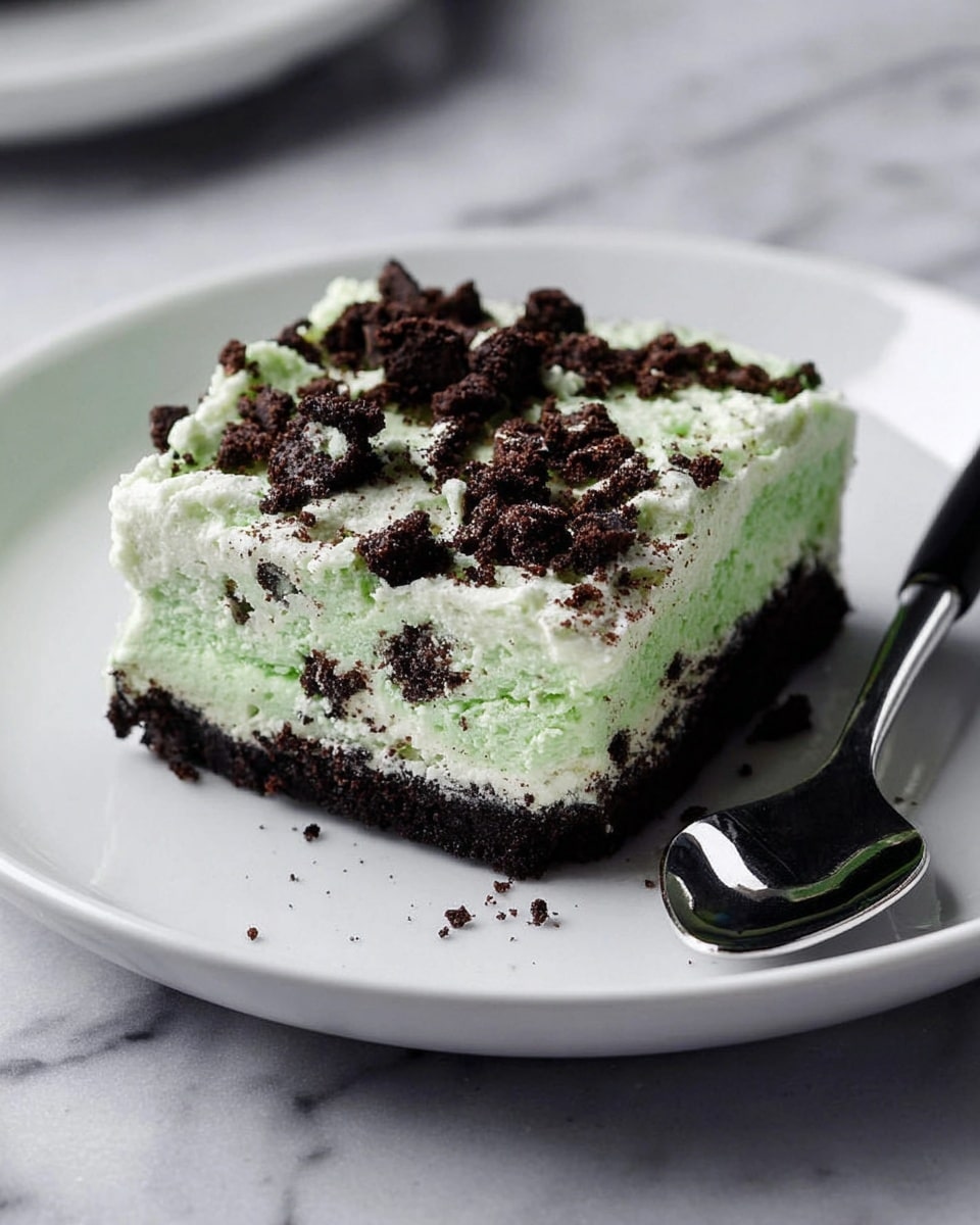 The image shows a square slice of a green creamy dessert with two main layers on a white plate. The bottom layer is dark, crumbly, and looks like crushed cookies forming a thin crust. The top layer is thick, light green, and fluffy with a smooth texture, sprinkled with uneven dark cookie pieces that add texture and contrast. A spoon with a shiny metal head and black handle is placed on the right side of the plate. The background is a white marbled texture. photo taken with an iphone --ar 4:5 --v 7