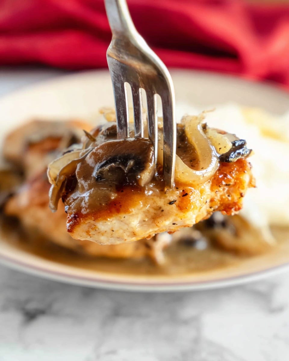 A close-up of a silver fork holding a piece of golden-brown cooked chicken with a slightly crispy texture on the edges, topped with light brown mushroom gravy containing small dark mushroom chunks and thin slices of cooked onions. The background shows a soft focus of a white plate and a red cloth, all on a white marbled surface. photo taken with an iphone --ar 4:5 --v 7