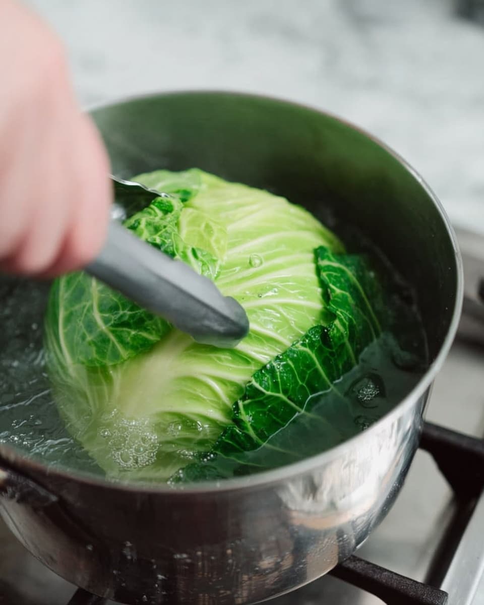 In the image, a large green cabbage is inside a silver pot filled with boiling water. The cabbage is vibrant green with some lighter green and white shades showing its leaf layers. A woman's hand is using silver tongs to hold the cabbage steady in the pot. The pot sits on a stove burner with a blurred white marbled surface around it. Water bubbles can be seen around the cabbage, showing the boiling process. The focus is on the cabbage and the silver pot, with soft natural light highlighting the textures. photo taken with an iphone --ar 4:5 --v 7