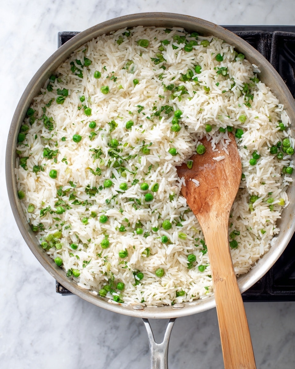 A large silver pan filled with cooked rice mixed with green peas and small chopped green vegetables evenly spread throughout. A wooden spoon rests inside the pan with some rice on its surface. The pan is placed on a black cooking stove, all over a white marbled surface. photo taken with an iphone --ar 4:5 --v 7