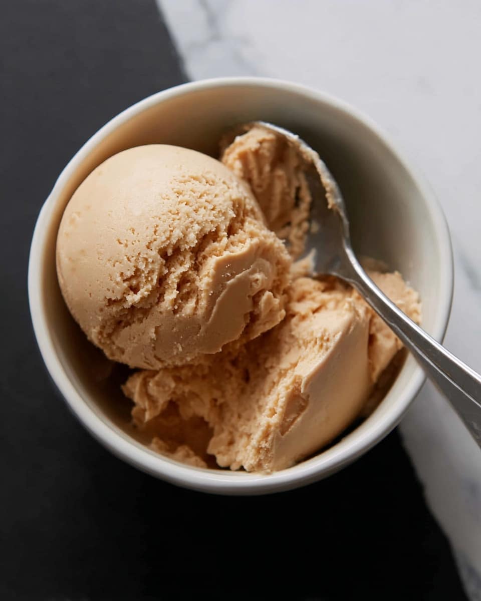 A round scoop of light brown ice cream sits in a small white bowl, showing a smooth and creamy texture with some rough edges. Next to the scoop inside the bowl is a silver spoon holding more of the same light brown ice cream, which looks soft and easy to scoop. The white bowl is placed on a surface with a white marbled texture, which adds a clean and elegant touch to the setting. photo taken with an iphone --ar 4:5 --v 7